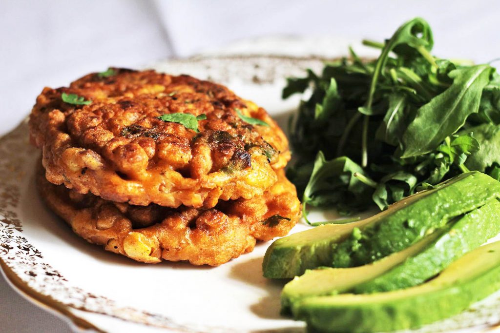 On a white plate with gold trim sits two sweetcorn and chickpeas fritters stacked along with a pile of rocket leaves and some sliced avocado