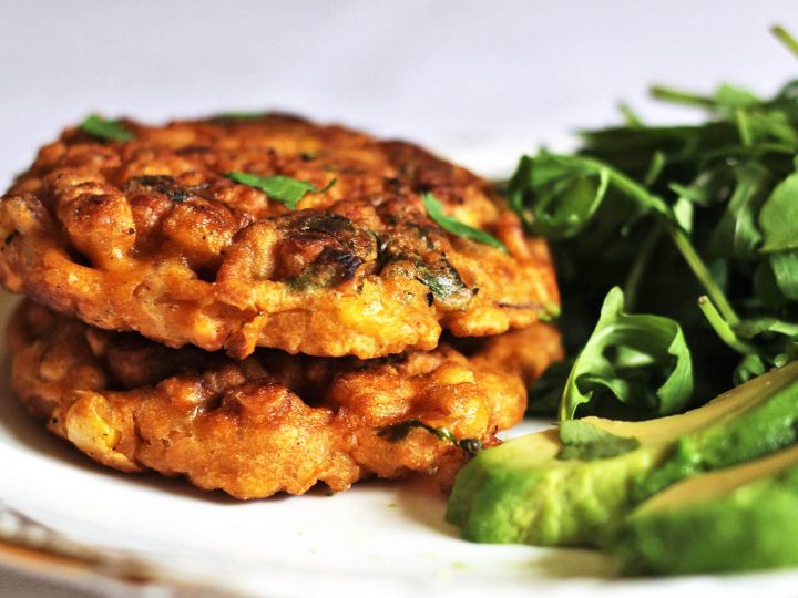 Two sweetcorn fritters stacked on a plate with a side of rocket leaves and sliced avocado