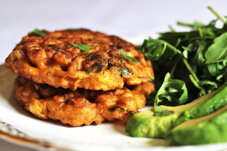 Two sweetcorn fritters stacked on a plate with a side of rocket leaves and sliced avocado